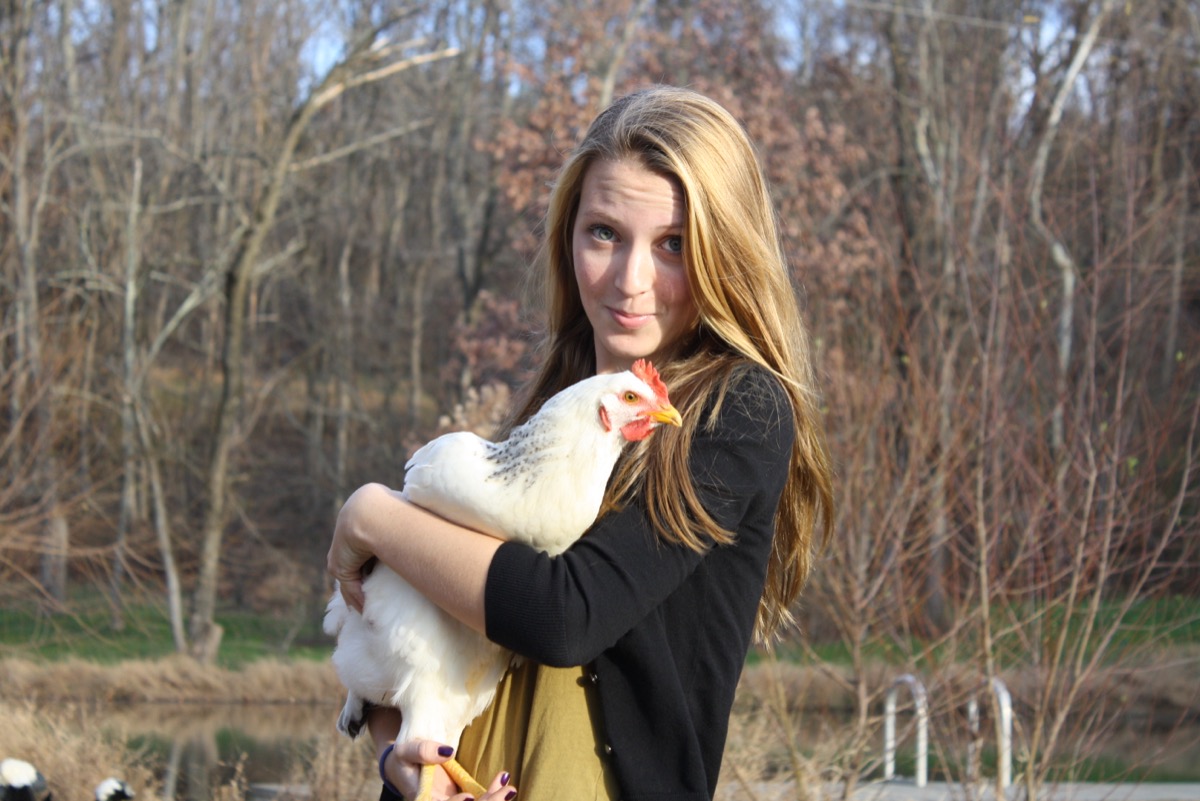 Finley smiling, holding a white chicken, with bare trees and a pond behind her